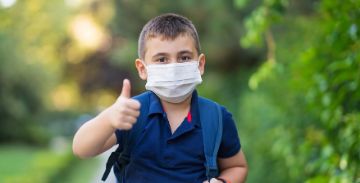 Male child wearing a white facemask and backpack giving a thumbs up to the camera, surrounded by greenery