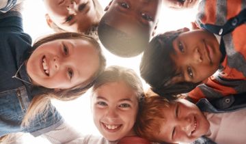 Diverse group of children in a circle leaning over, smiling, and looking down at the camera