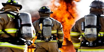 Back of three firefighters wearing fire gear with a large flame in the background