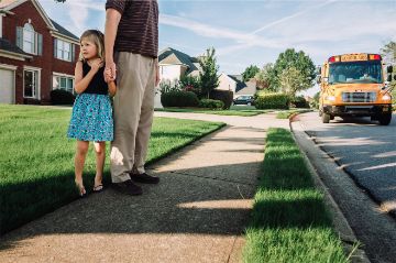 Father and daughter standing on the sidewalk in front of their house waiting for the school bus