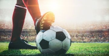 Legs standing on soccer field, one foot on the ball, stadium in the background