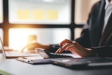 Woman in office wear working on laptop while using separate calculator with pen in hand