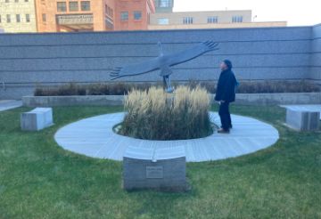A person stands on a concrete circle flanked by concrete benches in the middle of a lawn. He contemplated a sculpture of a bird flying with wings outstretched. The sculpture in its dark patina stands out against the straw-colored pampas grass the encircles the sculpture. 