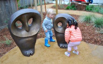 Two children of about 2-3 years in age play with one of two bronze sculptural owls. The owls have voids where the eyes are and one of the children, with black hair in ponytails, is peering through while a blond girl observes from the other side.