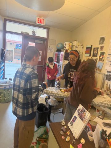 A teacher in a red hairband instructs students while working at an ironing board. The young teenagers are walking around an art classroom working on a project.
