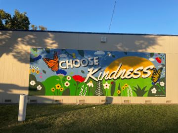 Butterflies, a ladybug and a bee fly through a garden of flowers with a bright sun in the background. Words reading "Choose Kindness" encourage love.