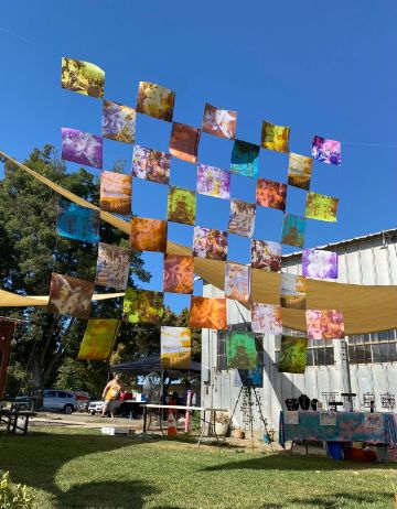 Many colorful paintings on fabric are stitched together to make a banner that hangs between two buildings.