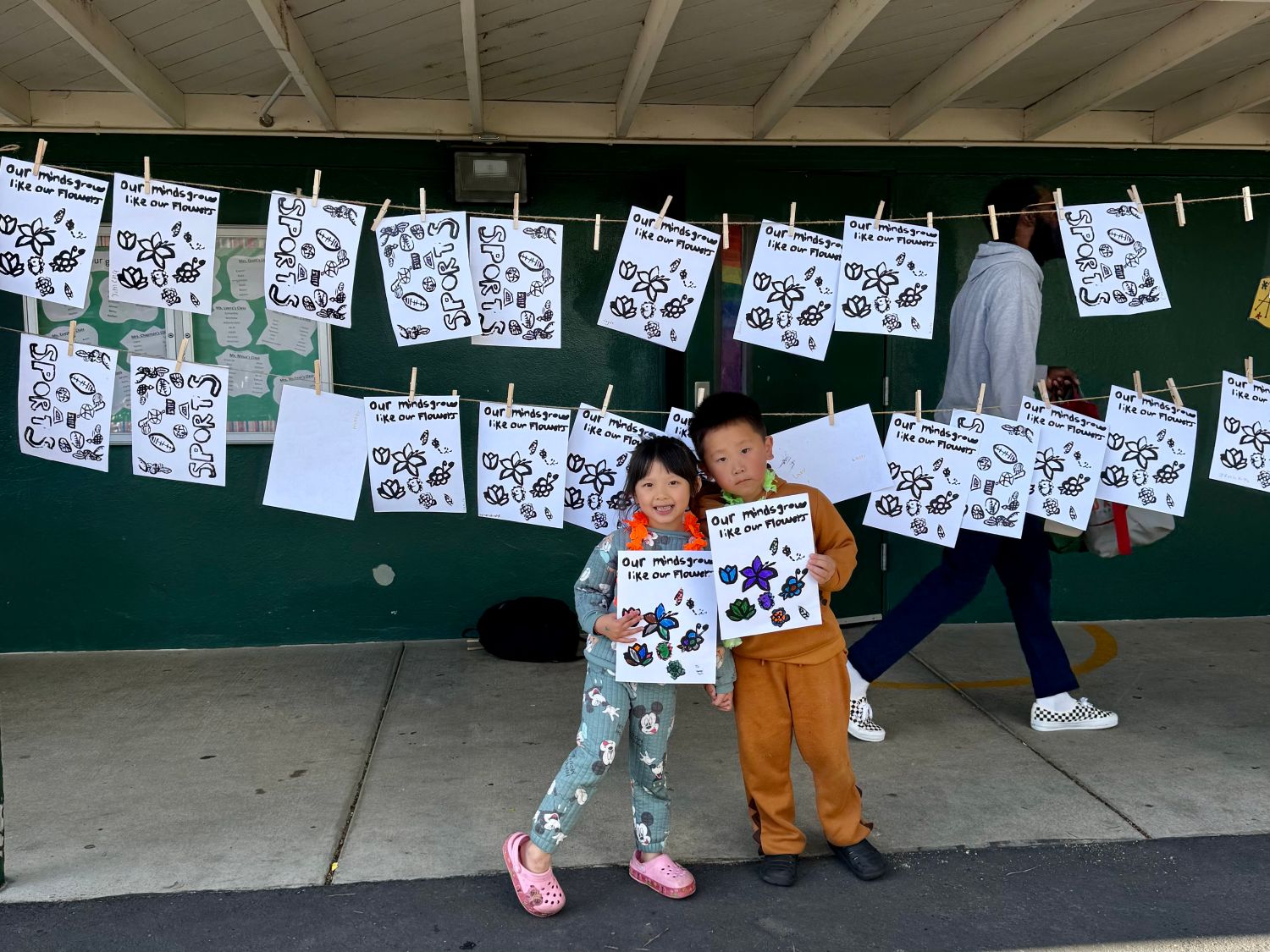 Two young children with big smiles, stand in front of a double clothesline that hangs heavy with prints. The students proudly present their own work.