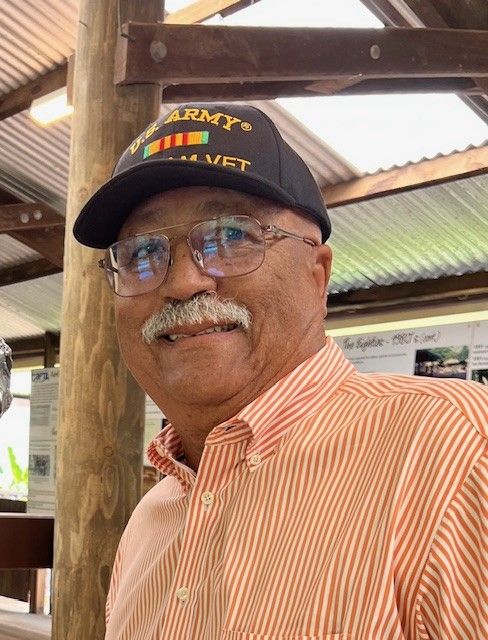A head-and-shoulders image of an older man. the man has a friendly smile and relaxed demeanor. He is wearing a peach dress shirt and a Viet Nam Vet's baseball cap.