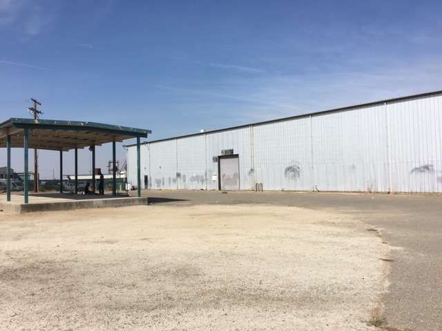 Grey building wall next to a shade structure against blue sky with a dirt road in the foreground.