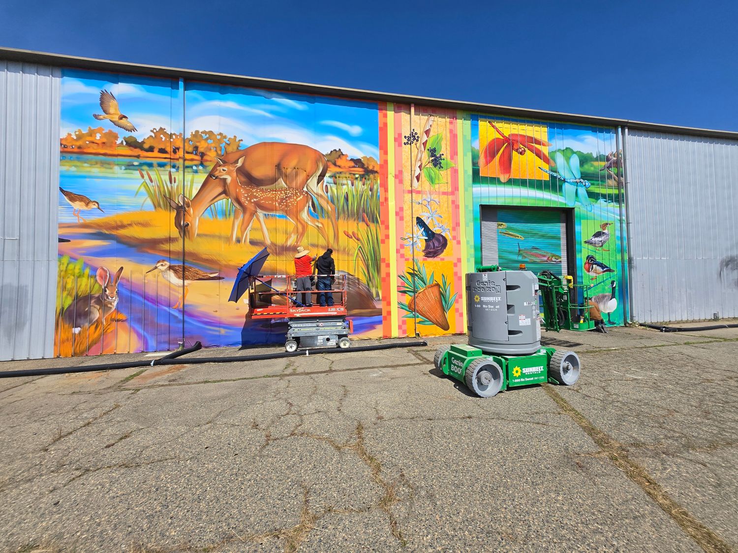 Vinyl mural being installed on to building next to a shade structure against blue sky with a dirt road in the foreground. 