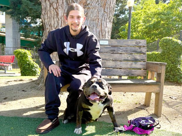 Jazz, who is a black and brindle Pit Bull,. sitting with her adopter on a wooden bench in a dog play yard