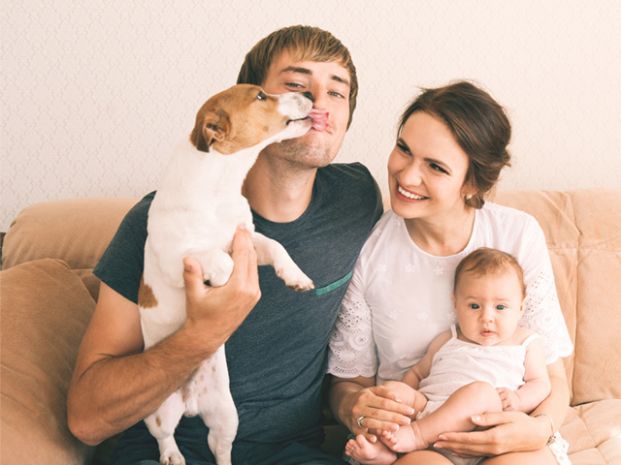 a small white dog being held up by a man sitting on a couch with a woman sitting alongside and holding a baby