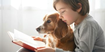 Young child reading a book with a dog in his lap in a sunny room