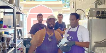 four men wear purple aprons cleaning dog bowls in the sink washing area