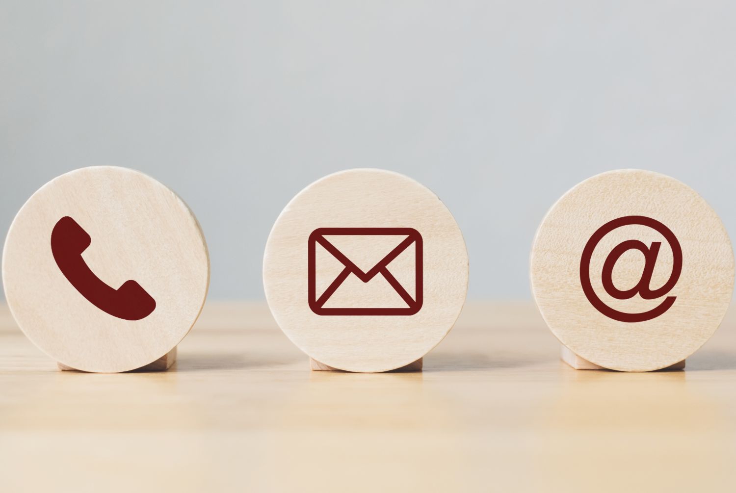 photo of three small round wooden blocks sitting on a wood base with images in dark brown from left to right, a phone, envelope and an @ sign. background is light grey.