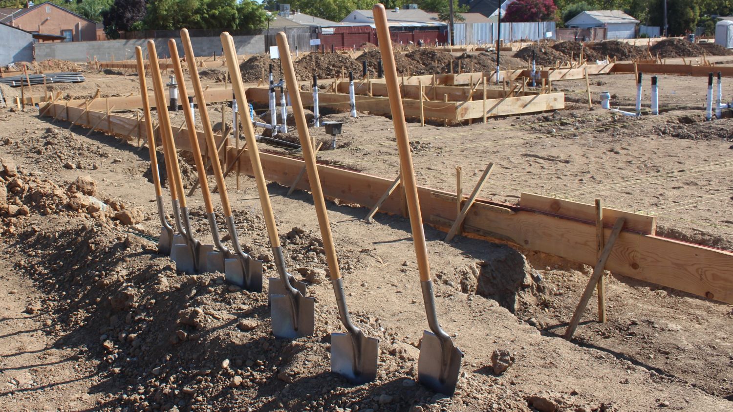 Row of shovels stuck upright in the dirt at a construction site, with wooden framing, soil piles, and early foundation work visible in the background.