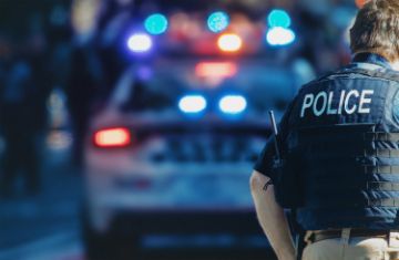 Police officer standing on the right in the foreground. Police car with lights on in the background.