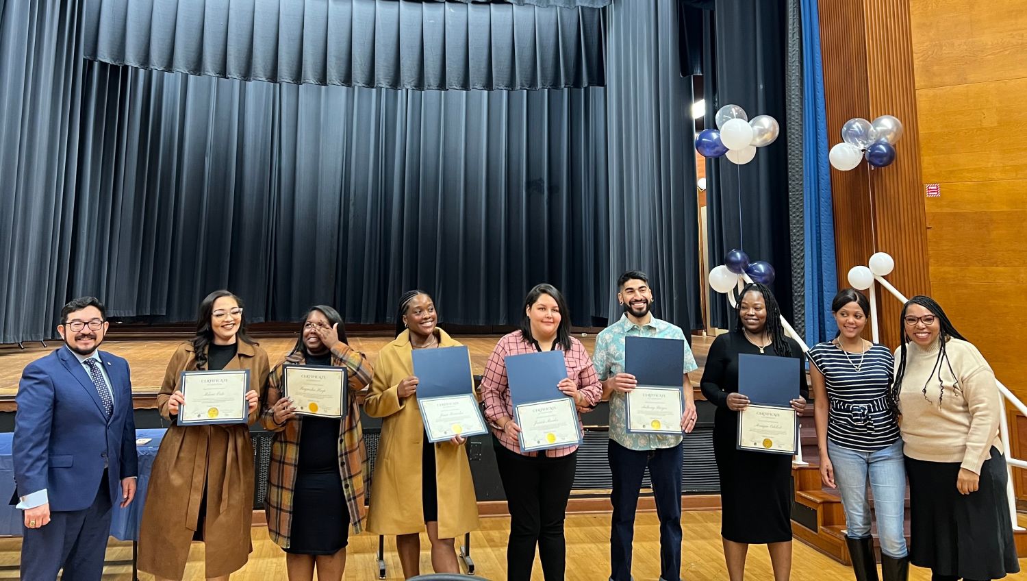 Six program graduates with certificates stand with program managers and city councilmember at graduation ceremony