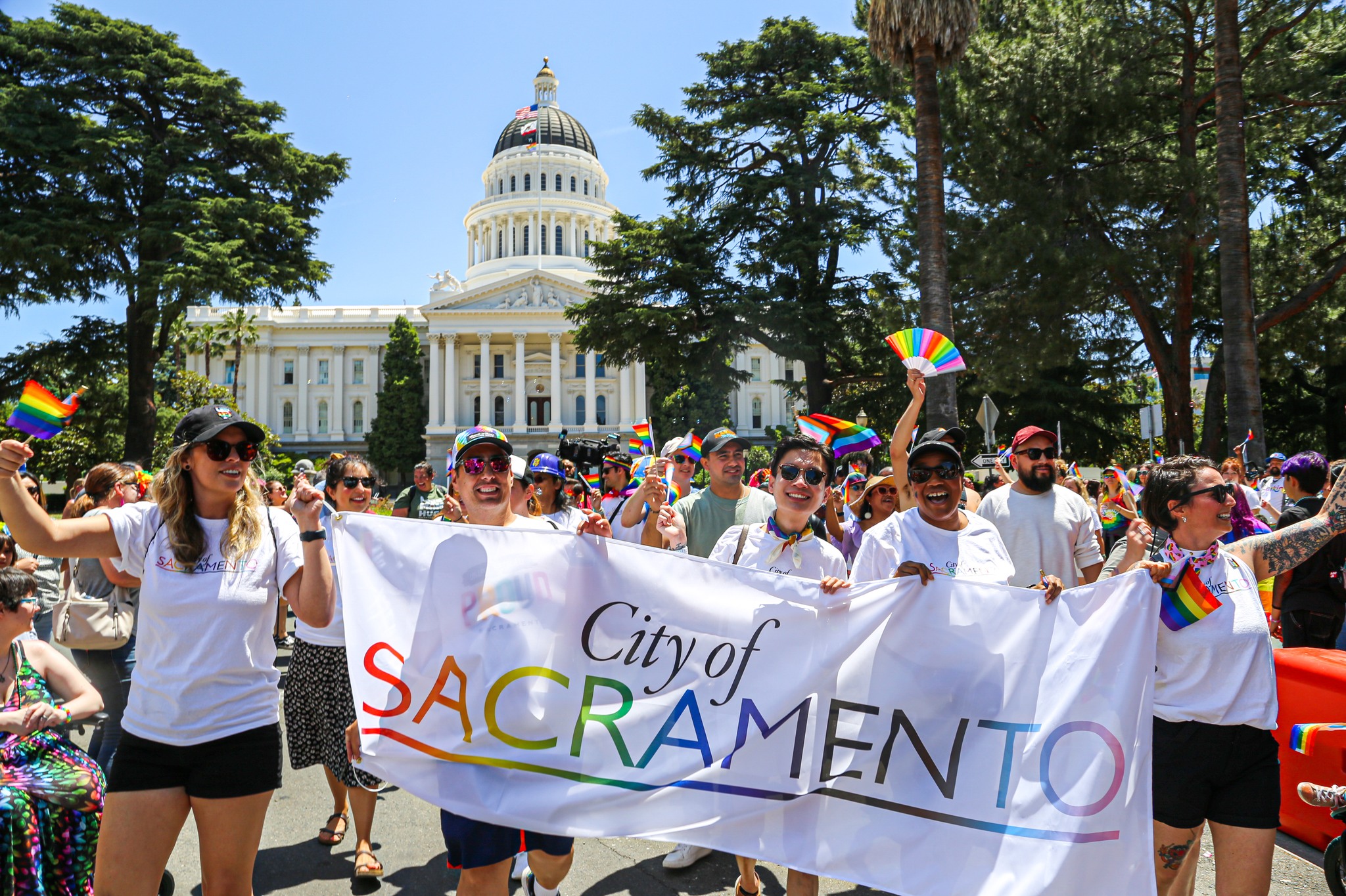 City employees stand in front of the CA capital building with a banner of the City of Sacramento pride logo.