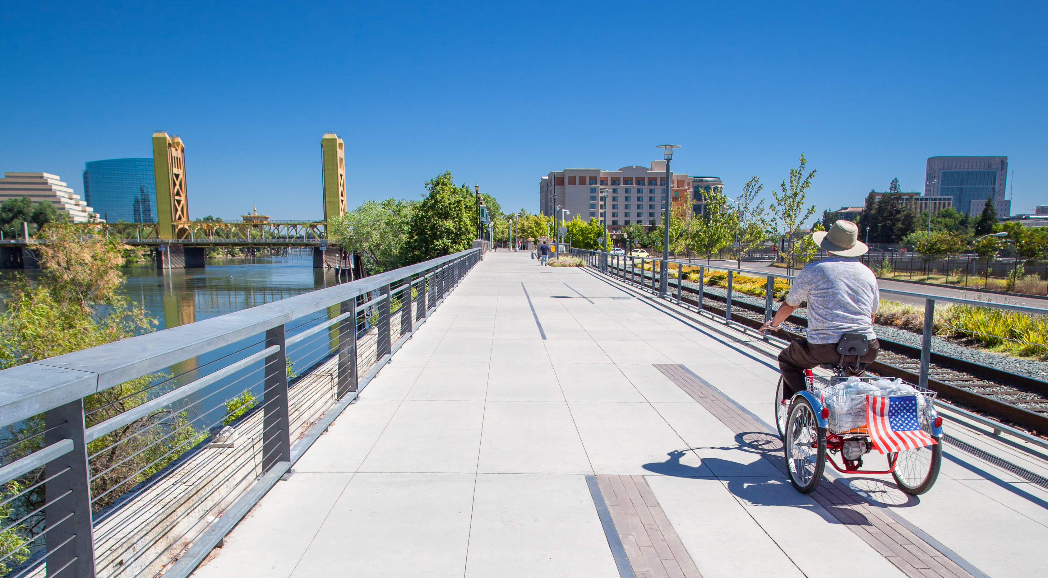 A person rides a three-wheel bike down a wide sidewalk near the Sacramento river. 
