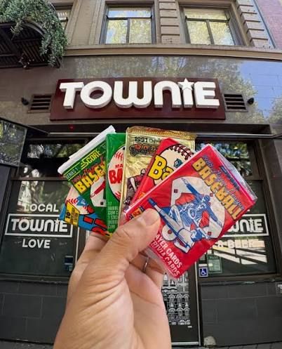 A hand holds packs of sports cards in front of the sign for the bar Townie, on K Street in Sacramento.
