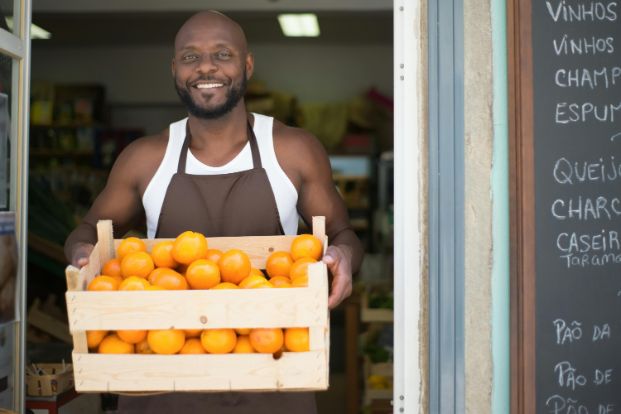 Image of a busienss owner in a doorway carrying  a crate of oranges. 