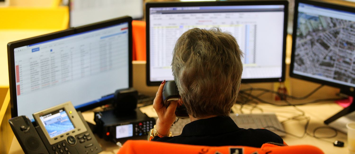 a 911 phone operator sitting in front of a desk with three computer monitors while on the phone.