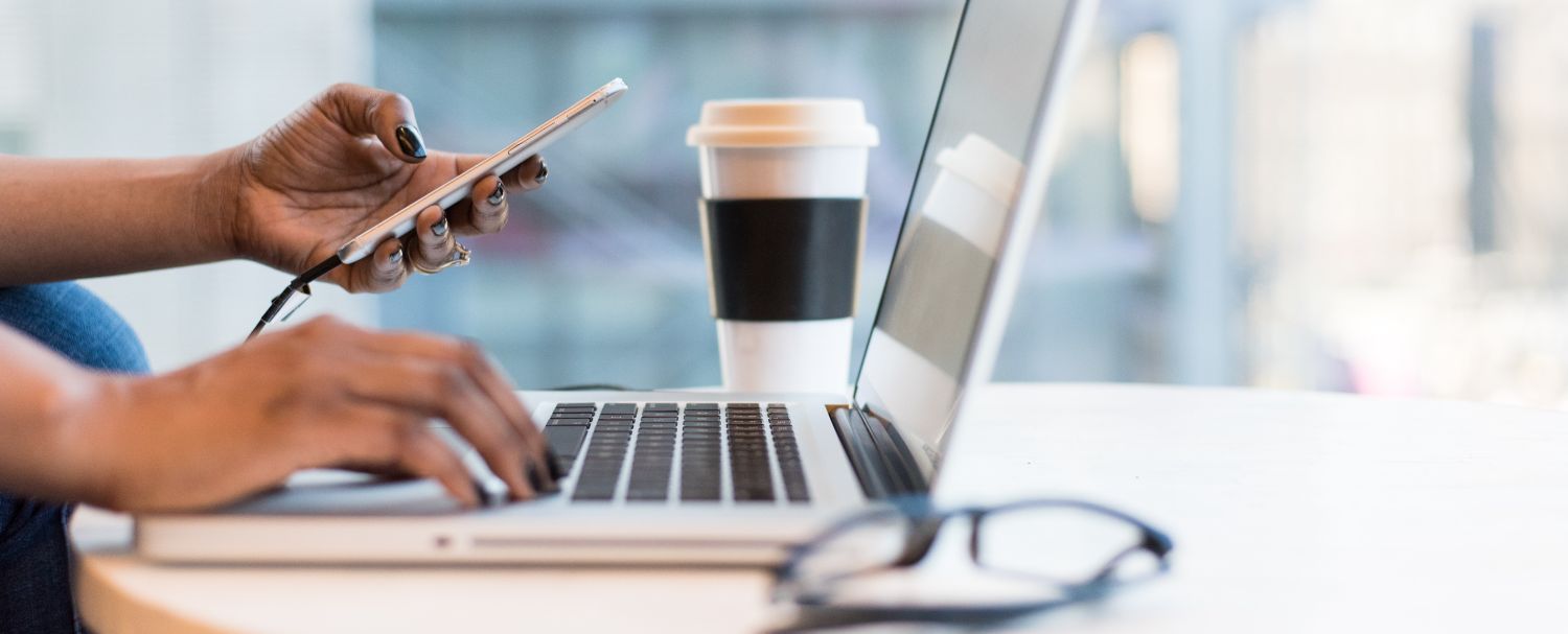 One hand typing on a laptop and the other holding a phone. In the background a cup of coffee and towards the front a pair of glasses.