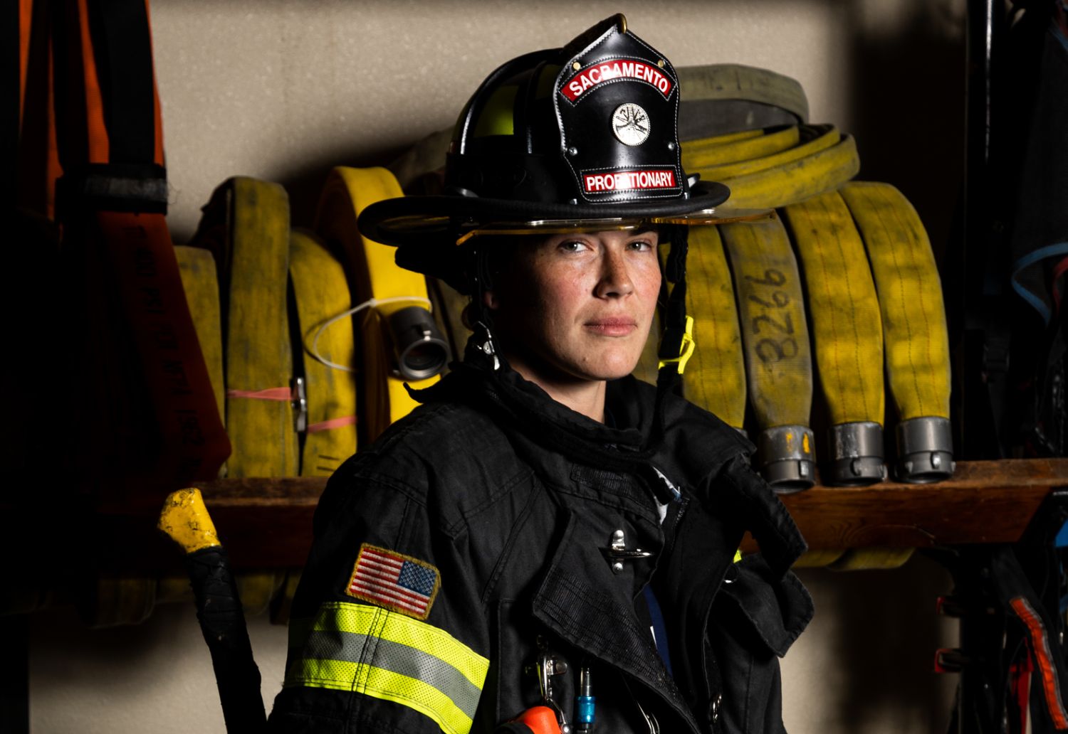 photo of a firefighter in full uniform with helmet that reads Sacramento Probationary. Yellow hose in the background. 