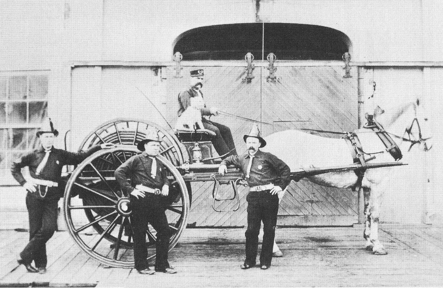 Black and white photo of four firefighters and a dog on a a horse and carriage.