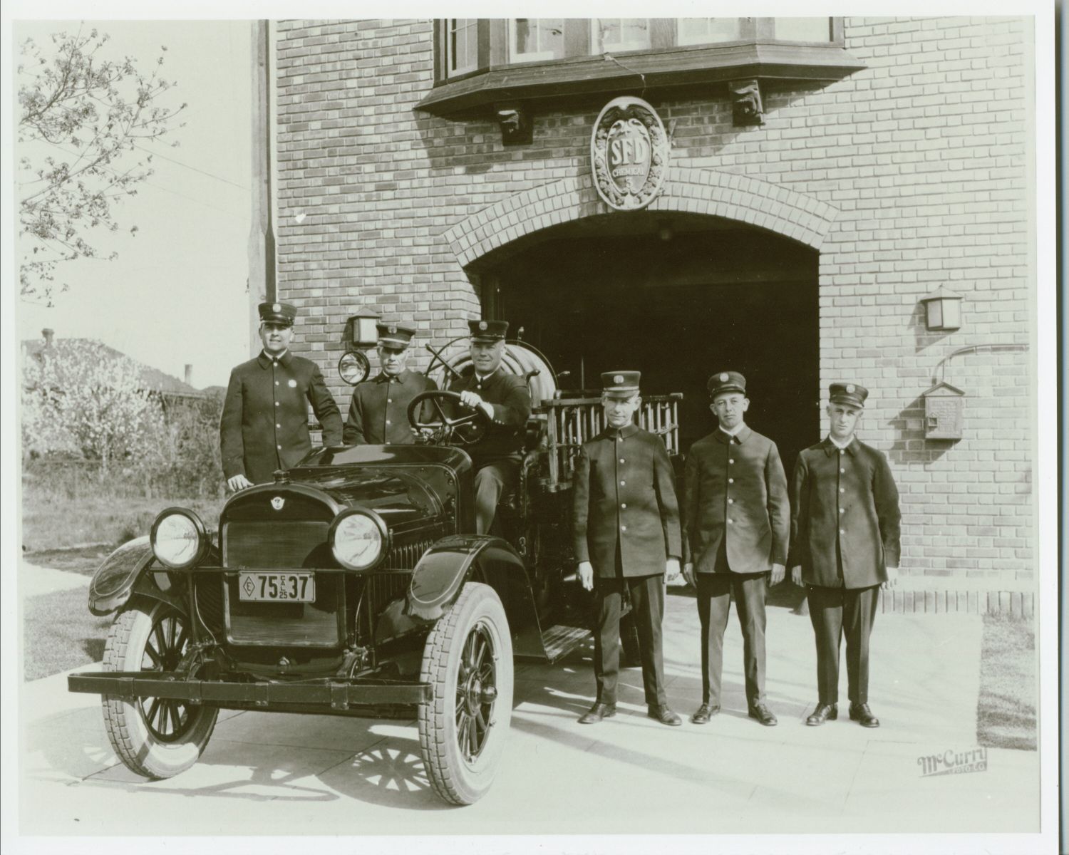 black and white photo of six firefighters standing with fire truck in front of a fire station.
