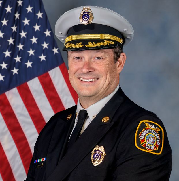 photo of assistant chief john danciart in full uniform with U.S. flag and gray background.