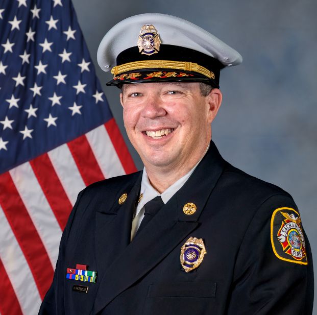 photo of fire chief costamagna in full uniform with hat. A gray background with U.S. flag.