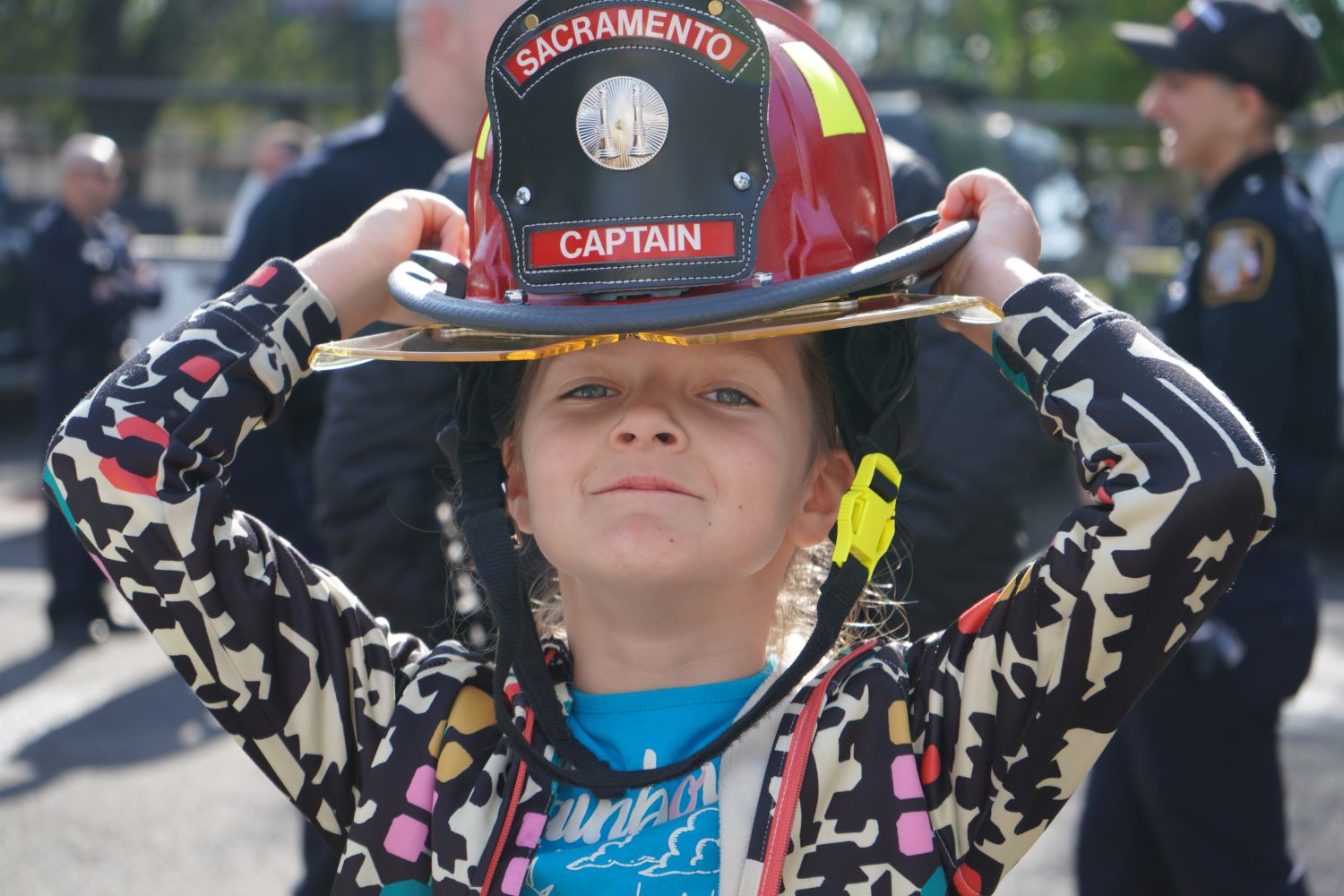 A YOUNG KID IN A COLORFUL SWEATER TRYING ON A FIRE CAPTAIN HELMET