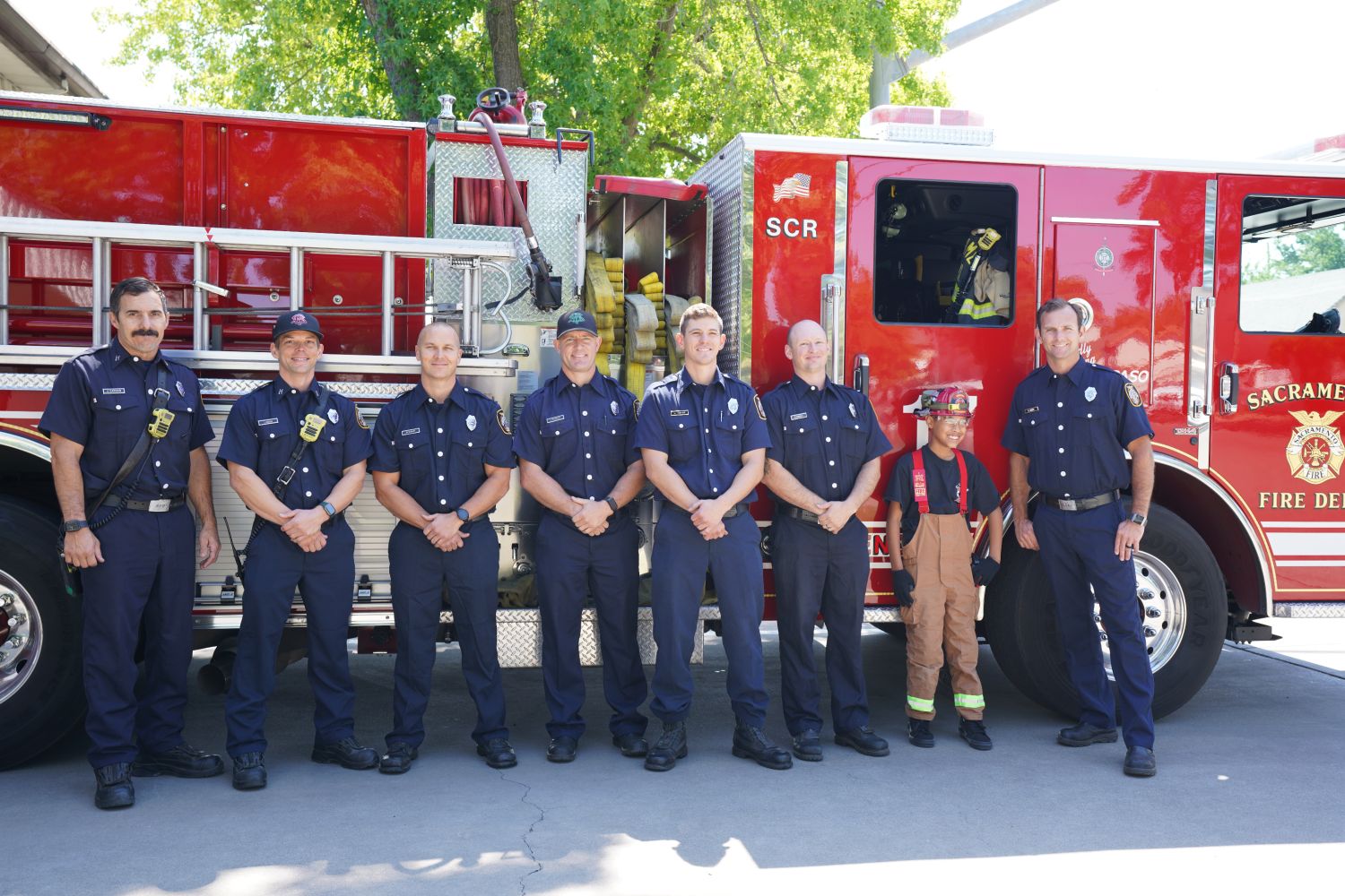 SEVEN FIREFIGHTERS LINED UP WITH A KID IN FRONT OF A FIRE ENGINE