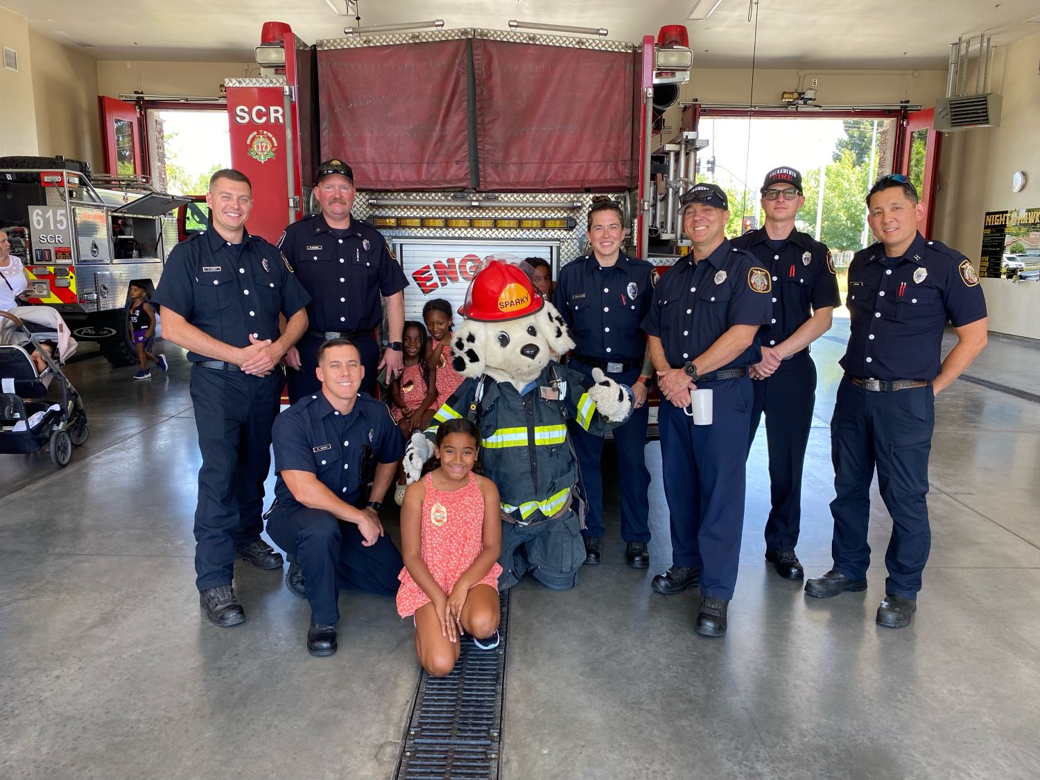 Seven firefighters posing with kids and the sparky mascot with a fire engine behind them.