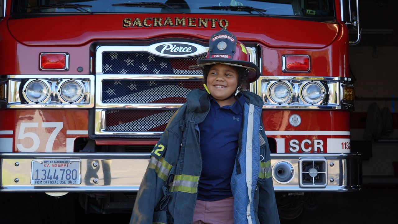 YOUNG BOY IN TURNOUTS AND FIRE HELMET STANDING IN FRONT OF A SAC FIRE ENGINE