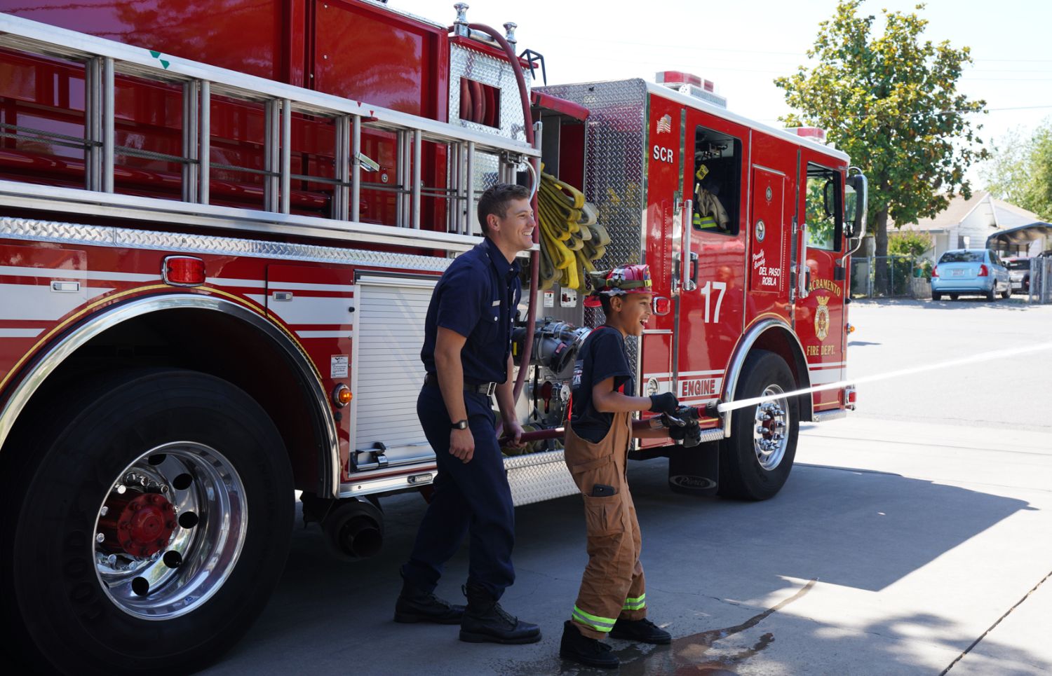 a young boy using a water hose with a firefighter assisting him, while standing in front of a fire engine.