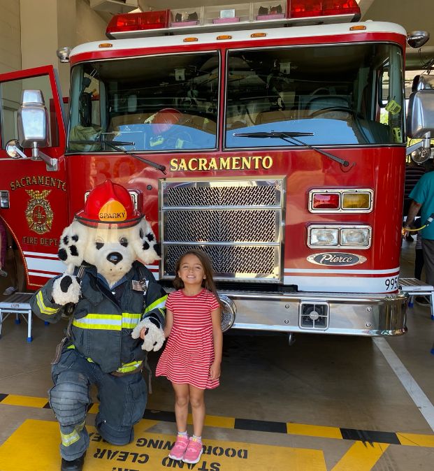 little girl standing next to sparky the fire mascot in front of the engine