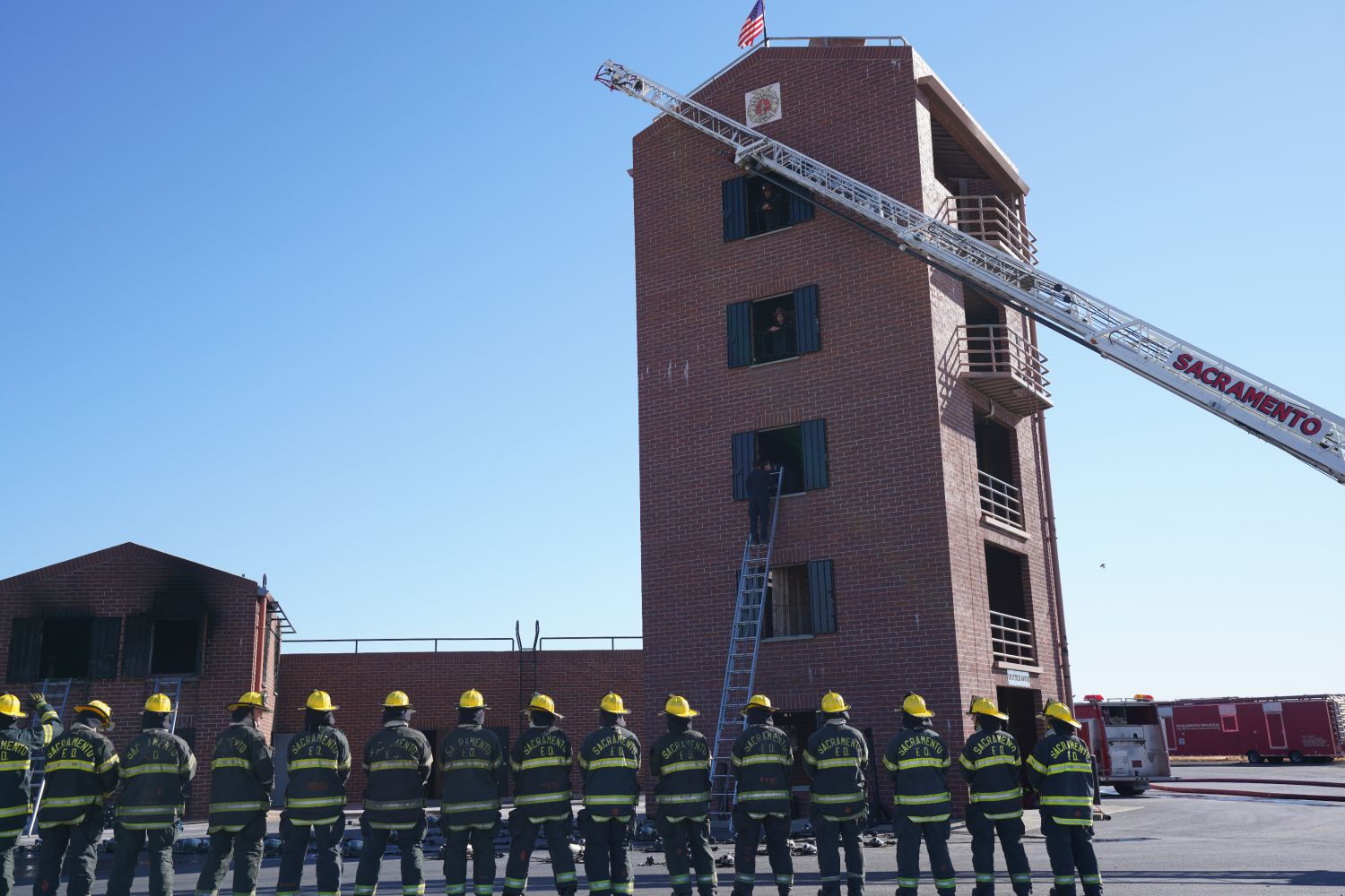 a line of firefighters at the training academy. A brick building with a ladder in front of them.
