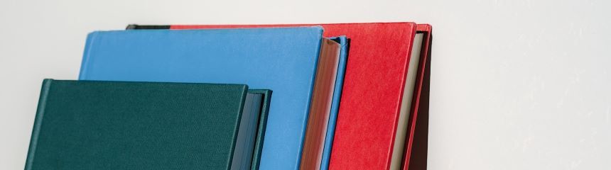 Three books  (greeen, blue and red) stacked atop each other, leaning against a wall