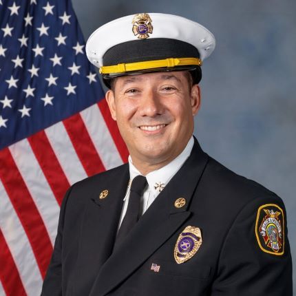 Fire Marshal Jason Lee standing in front of an American flag wearing a dark blue uniform with a Fire Marshal's badge and a white uniform cap