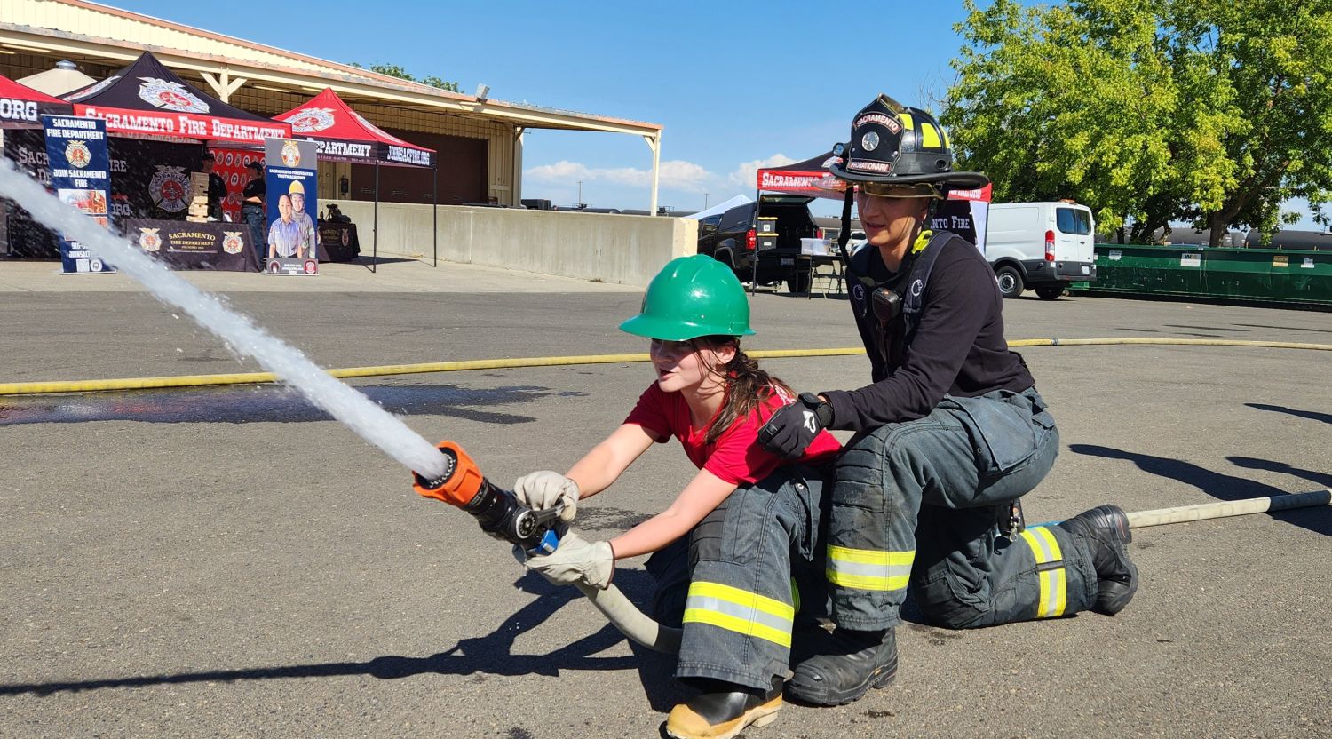 Camper kneeling on hose and being guided by a firefighter in black how to squirt water out of a fire hose