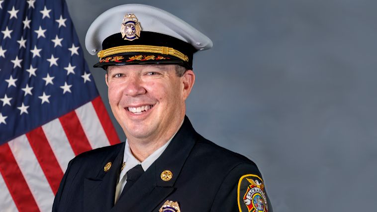 photo of Sacramento Fire Chief in uniform with hat. The background is gray with U.S. flag. 