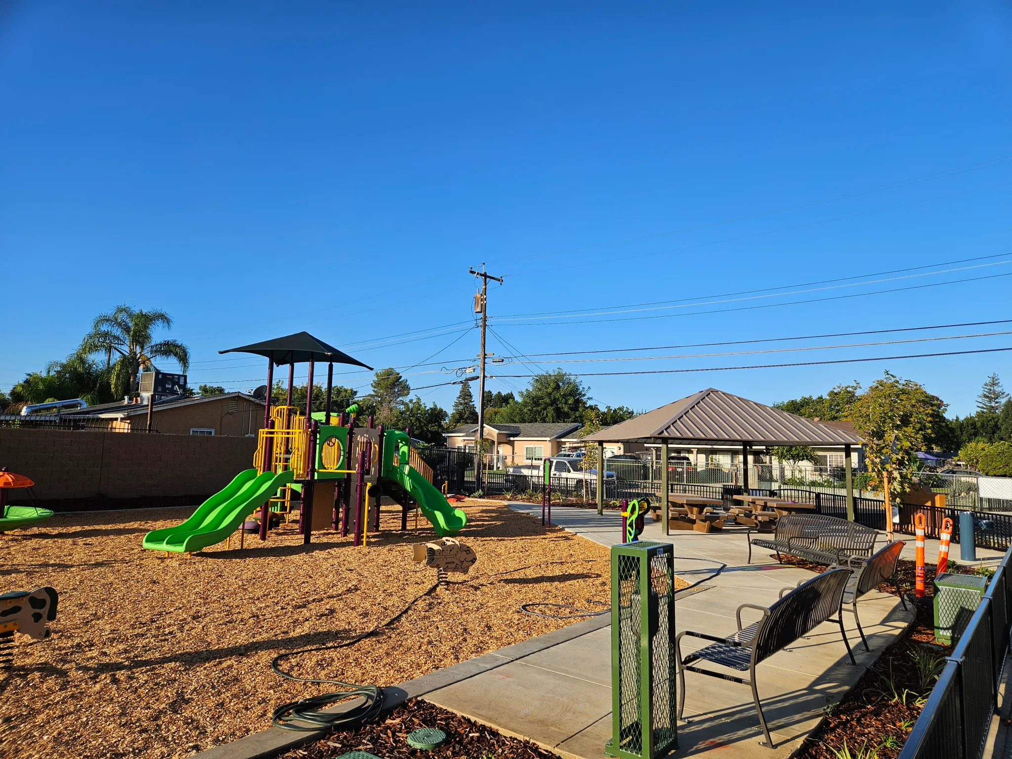 A bright green and yellow playground with slides sits on wood chips next to benches and a covered picnic area under a clear blue sky in a Sacramento neighborhood park.