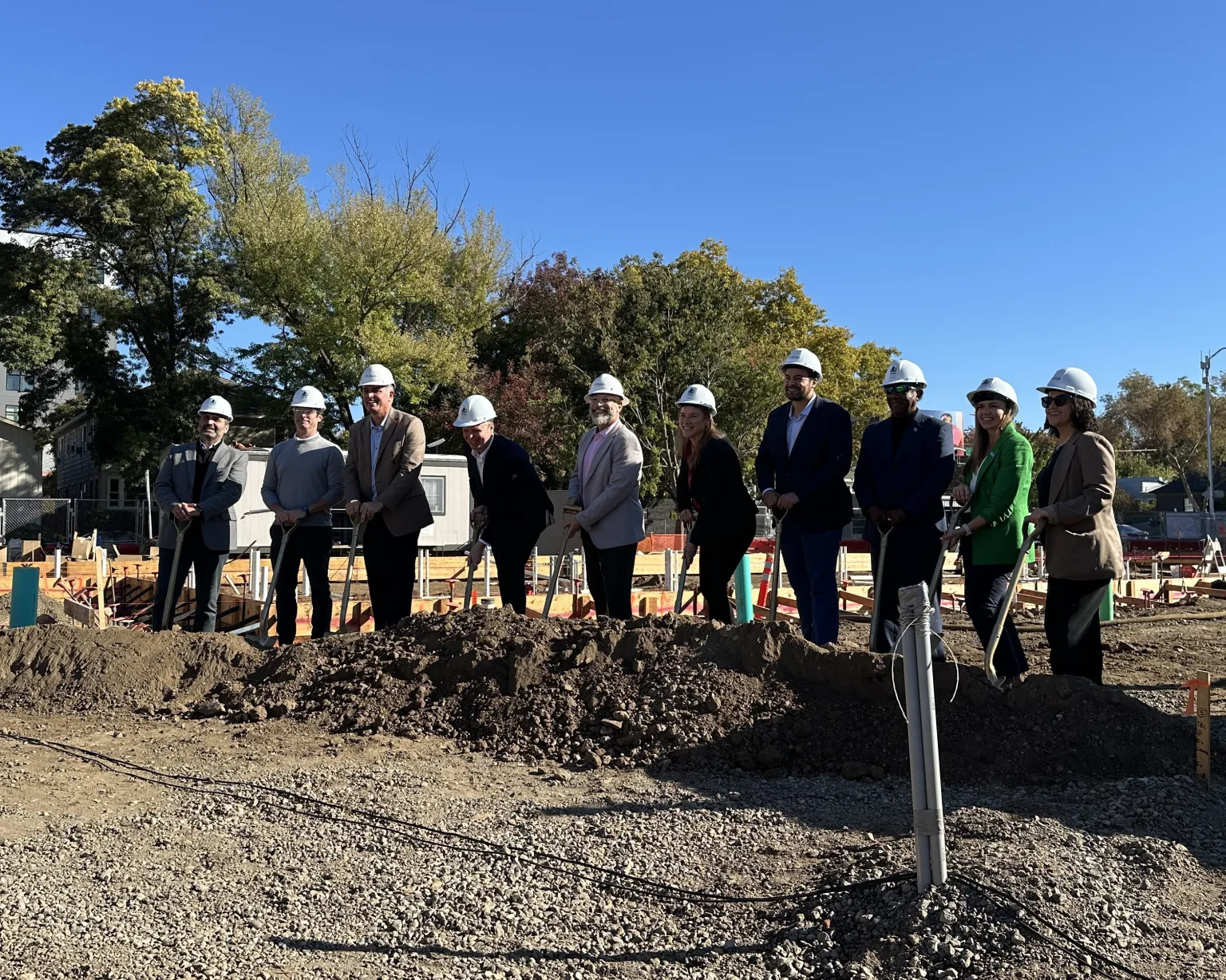 Ten people wearing white construction helmets stand together, holding shovels at a groundbreaking ceremony.