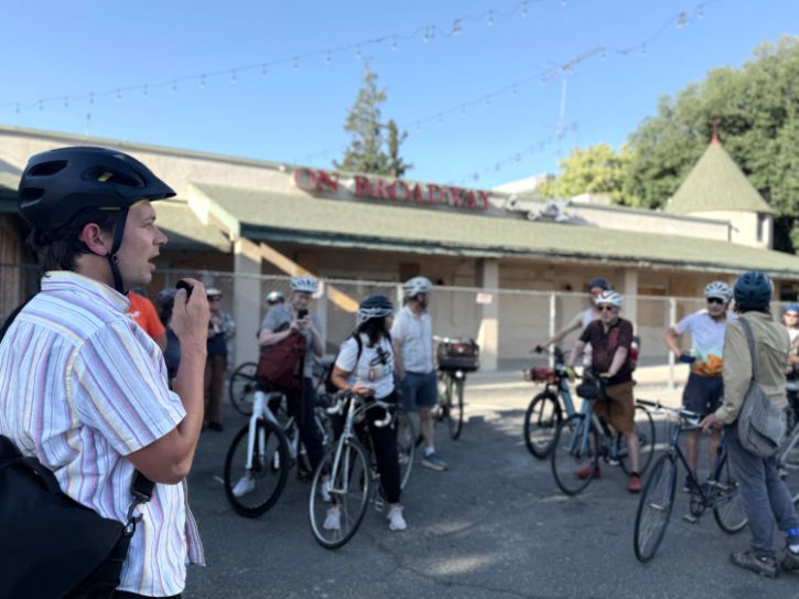 Person speaking into microphone to a group of people on bikes wearing helmets.