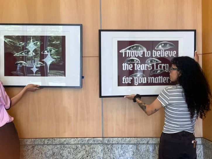 Two artists hanging framed artwork on wall of exhibit.