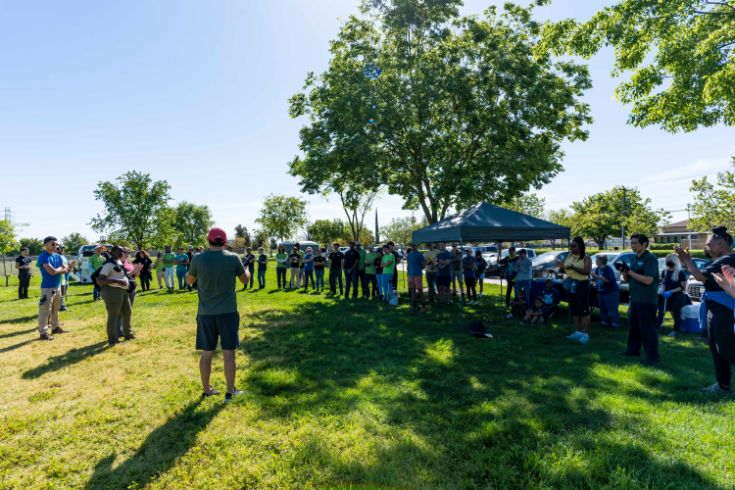 Large group of people gathered in field at a park. Trees, tent canopy